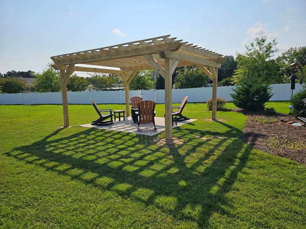 Wooden pergola with Adirondack chairs on a grassy lawn, casting shadows under bright blue sky.