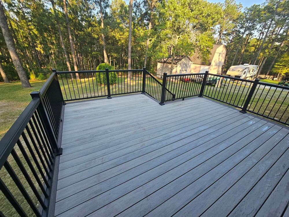 Spacious outdoor deck with black railing surrounded by trees and a house in the background.